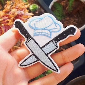 Può includere: Toppa ricamata bianca con due coltelli incrociati e un cappello da chef blu. I coltelli sono neri con lame argentate.