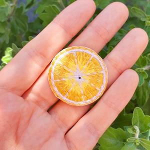 Könnte beinhalten: Eine Hand hält eine kleine gestickte Orangenscheibe. Die Orangenscheibe ist gelb mit einer weißen Mitte und einem braunen Stiel.