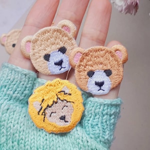 Könnte beinhalten: Drei gestickte Aufnäher mit einem Bären, einem Bären und einem Löwen. Die Aufnäher sind beige, braun und gelb. Die Aufnäher befinden sich auf einer Hand, die einen hellblauen Strickhandschuh trägt.