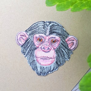 May include: Embroidered patch of a chimpanzee's face with black fur, pink nose, and brown eyes. The patch is on a brown background.