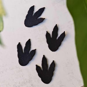 May include: Four black dinosaur footprint cutouts arranged on a white background. Each footprint has three toes. The cutouts are made of a textured material, and the image is well-lit, highlighting the details of the footprints.