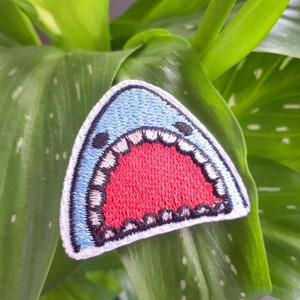 Peut inclure: Un patch brodé de requin bleu et rouge avec un contour blanc et une grande bouche ouverte. Le patch est sur un fond de feuilles vertes.