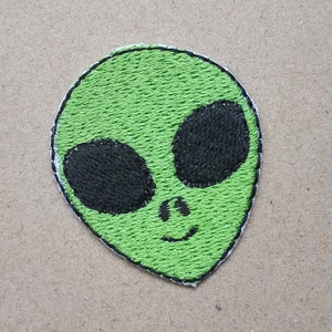 Könnte beinhalten: Grüne Alien-Stickerei mit schwarzen Augen und einem lächelnden Mund.