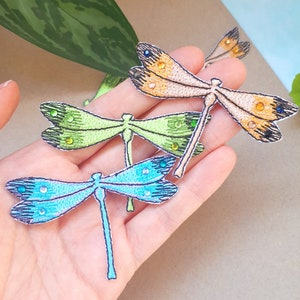 Könnte beinhalten: Drei gestickte Libellen-Patches in verschiedenen Farben: Blau, Grün und Orange. Jeder Patch hat einen perlenbesetzten Körper und Flügel mit einem detaillierten Design.