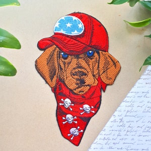 Könnte beinhalten: Gestickter Patch eines Hundes, der eine rote Baseballkappe mit einem blauen und weißen Sternenmuster und ein rotes Bandana mit weißen Totenköpfen und Knochen trägt.