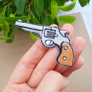 Könnte beinhalten: Ein gestickter Bügeleisen-Aufnäher mit einem silbernen Revolver und einem orangefarbenen Griff.