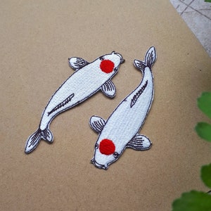 Könnte beinhalten: Zwei weiße Koi-Fisch-Aufnäher mit schwarzen Umrissen und einem roten Kreis auf jedem. Die Aufnäher sind gestickt und liegen auf einer braunen Oberfläche. Ein dekorativer Artikel.