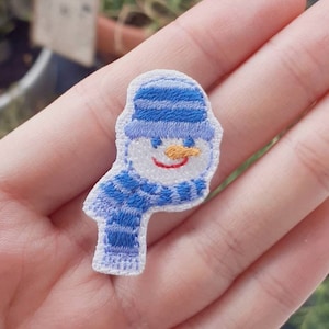 Könnte beinhalten: Ein weißer Filz-Schneemann mit einer blau-weiß gestreiften Mütze und einem Schal. Der Schneemann hat eine Karottennase, schwarze Augen und ein rotes Lächeln.