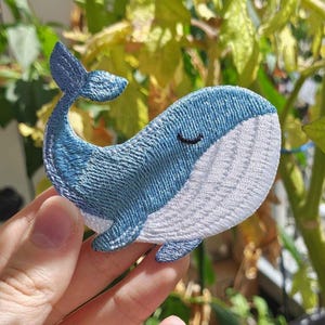Könnte beinhalten: Ein gestickter Aufnäher in Form eines Wals. Der Wal hat einen blauen Oberkörper und einen weißen Bauch mit horizontalen Sticklinien. Er besitzt eine kleine, geschwungene schwarze Linie als geschlossenes Auge und eine hochstehende Schwanzflosse.