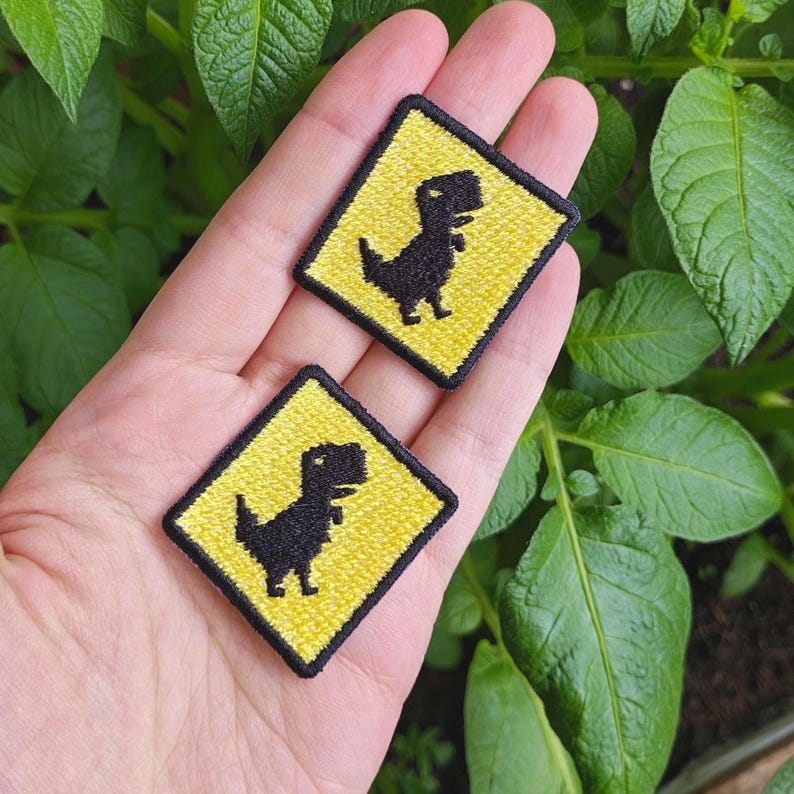 May include: Two yellow and black embroidered iron-on patches featuring a pixelated dinosaur in a warning sign shape.