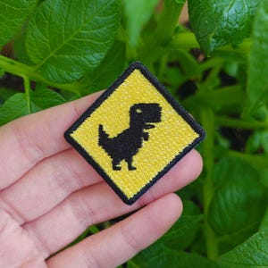 Può includere: Un cartello di avvertimento giallo a forma di diamante con un dinosauro pixelato nero al suo interno. Il cartello è ricamato con una superficie testurizzata.