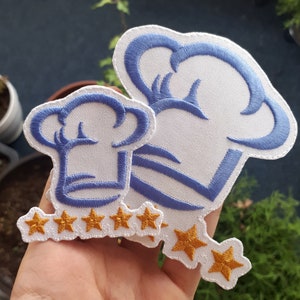 Può includere: Due cappelli da chef in feltro bianco con dettagli ricamati blu e cinque toppe a stella dorate. I cappelli sono cuciti insieme con le stelle in mezzo.