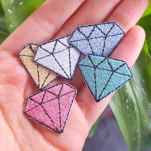 Könnte beinhalten: Fünf gestickte Diamanten-Patches in verschiedenen Farben, darunter Pink, Blau, Grün und Gold, auf einer Hand.
