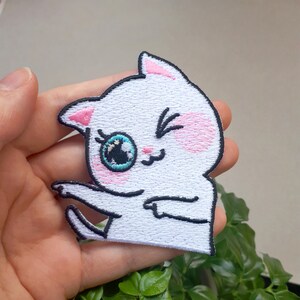 Könnte beinhalten: Weißer gestickter Aufnäher mit einem Cartoon-Katzenmotiv, das ein Auge geschlossen hat, zwinkert und einen rosa Rouge auf den Wangen hat. Die Katze ist weiß mit schwarzen Umrissen und blauen Augen.