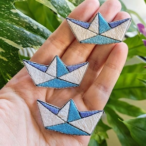 Könnte beinhalten: Drei gestickte Aufnäher in Form von Papierbooten. Jeder Aufnäher hat ein weißes, hellblaues und dunkelblaues Design mit schwarzen Nähten. Die Aufnäher werden auf einer Hand präsentiert, mit grünem Laub im Hintergrund.