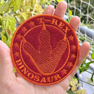 May include: Circular embroidered patch featuring a T-Rex footprint design. The patch has a red base with orange stitching and the words "T-REX DINOSAUR". The patch is approximately 7.6 cm in diameter.