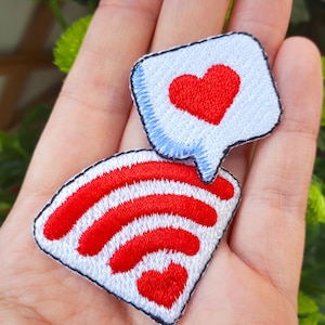 Puede incluir: Dos parches bordados para planchar. Uno es un parche blanco con un símbolo de wifi rojo. El otro es un parche azul con un globo de diálogo blanco y un corazón rojo dentro.