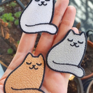 Op de afbeelding: Drie geborduurde strijkpatches van katten in verschillende kleuren: wit, grijs en oranje. Elke kat zit met zijn ogen dicht en een kleine glimlach.