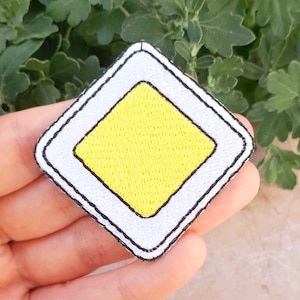 Könnte beinhalten: Gestickter Aufnäher mit einer gelben Raute in einem weißen Quadrat, umrandet von Schwarz. Der Aufnäher ist ein Verkehrsschild, das eine Vorfahrtsstraße kennzeichnet. Der Aufnäher wird in einer Hand gehalten.
