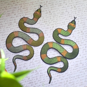 Könnte beinhalten: Zwei gestickte Schlangen-Patches in Grün und Orange auf weißem Hintergrund mit handschriftlichem Text.
