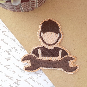 Puede incluir: Parche de planchado bordado marrón de un mecánico de dibujos animados sosteniendo una llave inglesa. El mecánico lleva una gorra y un mono marrones.