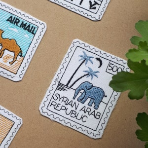 Könnte beinhalten: Ein weißer Stempel aus Stoff mit schwarzem Rand und einem blauen Elefanten, der unter zwei Palmen steht. Der Stempel trägt die Aufschrift "SYRIAN ARAB REPUBLIC" und "500" in schwarzer Schrift.