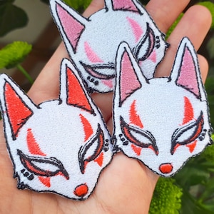 May include: Embroidered patches featuring a stylized fox mask design. The masks have white faces with red and pink accents, black details, and a red nose. The patches are likely for clothing or accessories.
