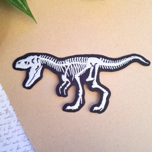 Könnte beinhalten: Weiß gestickter Dinosaurierskelett-Patch auf schwarzem Hintergrund.