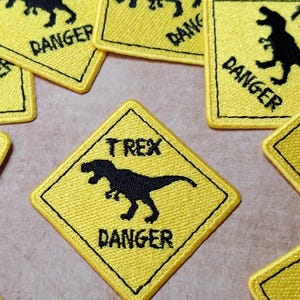 May include: Yellow and black embroidered patches with a dinosaur silhouette and the words "TREX DANGER". The patches are square with a black border and a diamond shape. The patches are scattered on a neutral surface.