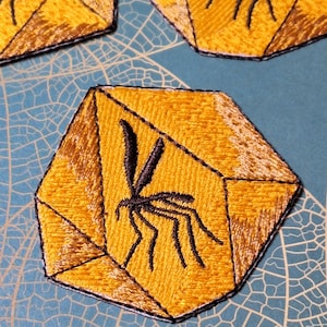 Puede incluir: Parches bordados con un diseño geométrico amarillo y una silueta de insecto negro. Los parches tienen un borde negro y están colocados sobre un fondo azul claro con un patrón similar a una hoja.