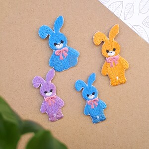 May include: Four embroidered bunny patches in different colors: blue, purple, orange, and light blue. Each bunny has a pink bow and a white face.