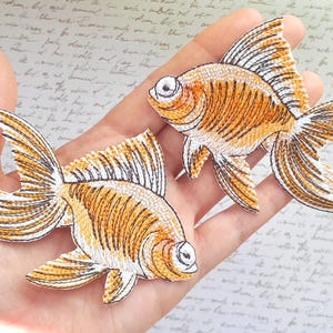 Może przedstawiać: Dwa haftowane naszywki z motywem złotych rybek. Ryby są pomarańczowe i białe z czarnymi konturami. Naszywki idealnie nadają się do dodania odrobiny fantazji do ubrań, toreb lub innych przedmiotów z tkaniny.