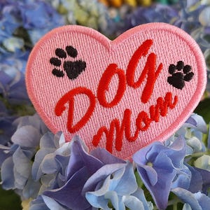 Könnte beinhalten: Ein rosa herzförmiger, gestickter Aufnäher mit den Worten "Dog Mom" in roter Schrift und zwei schwarzen Pfotenabdrücken. Der Aufnäher befindet sich vor einem Hintergrund aus blauen und violetten Hortensienblüten.