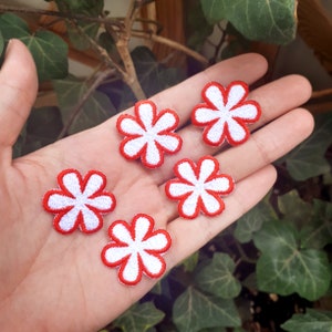 Può includere: Cinque toppe ricamate a forma di fiore, con petali bianchi e contorni rossi. Le toppe hanno la forma di un fiore a sei petali. Le toppe sono piccole e sembrano essere fatte di tessuto. Sono tenute in una mano.