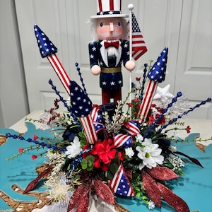 May include: A patriotic centerpiece featuring an Uncle Sam nutcracker, American flags, and star-spangled rockets. The arrangement includes red, white, and blue floral accents, all set on a turquoise decorative base. A festive display for Independence Day.