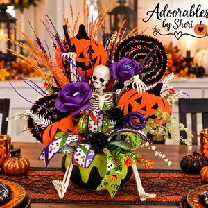 May include: A Halloween centerpiece featuring a skeleton figure in a lime green cauldron, accented with orange felt pumpkins, purple roses with eye centers, black glittered swirls, and a multi-colored polka dot ribbon. Text reads: Adorables by Sheri.