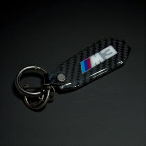 May include: A carbon fiber key chain with a silver clasp and key rings. The key chain features the BMW M logo in blue, red, and white. The background is black.