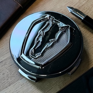 Puede incluir: Un emblema cromado brillante con un diseño detallado de cabeza de caballo. El emblema es circular con un perfil de caballo en relieve y texturizado sobre un fondo oscuro. Un bolígrafo negro es visible en el fondo.