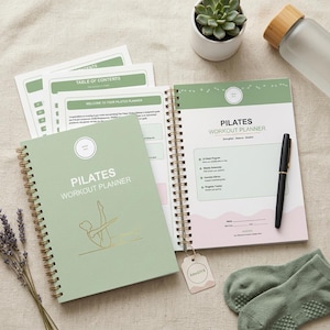 Peut inclure: Un planificateur d'entraînement Pilates avec une couverture vert sauge et une reliure spirale dorée. Le planificateur comprend un programme de 12 semaines, des horaires hebdomadaires et un suivi des progrès. Un stylo noir, une bouteille d'eau et des chaussettes sont également visibles.