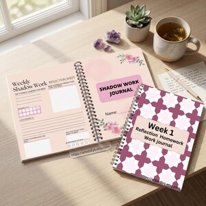 Peut inclure: Deux journaux à spirales avec des couvertures roses et des accents floraux. L'un des journaux est ouvert, affichant le texte "Weekly Shadow Work" et "Reflection Sheet". L'autre journal est fermé, avec le texte "Week 1 Reflection Homework Work journal". Une tasse de thé et une plante succulente sont également visibles.