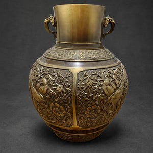 May include: A metallic bronze-toned vase featuring intricate relief carvings of birds and floral patterns across its rounded body. The vessel includes two small handles shaped like animal heads on the neck and a smooth, polished rim at the top.