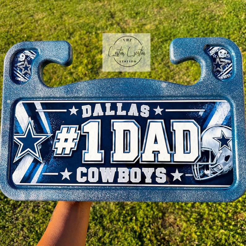 May include: A rectangular blue glitter car headrest tray featuring a Dallas Cowboys theme. The center graphic displays the text DALLAS #1 DAD COWBOYS with a star logo and a football helmet. The tray includes two hooks for mounting on seat posts.