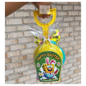 May include: A yellow plastic shovel-shaped Easter basket filled with gifts, wrapped in cellophane, and tied with a decorative bow. The basket features a "Happy Easter" graphic with SpongeBob SquarePants and Easter eggs.