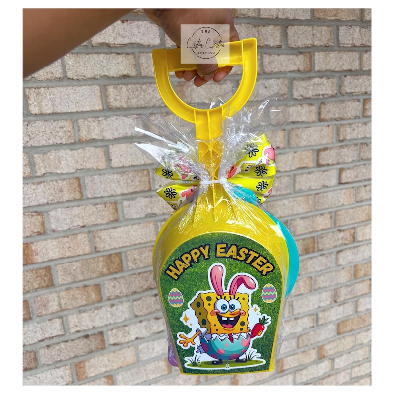 May include: A yellow plastic shovel-shaped Easter basket filled with gifts, wrapped in cellophane, and tied with a decorative bow. The basket features a "Happy Easter" graphic with SpongeBob SquarePants and Easter eggs.