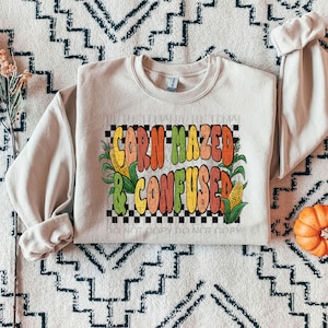 May include: A beige sweatshirt with a graphic print of a checkered background with the text "Corn Mazed & Confused" in colorful, retro-style lettering. The graphic also features corn stalks and ears of corn.