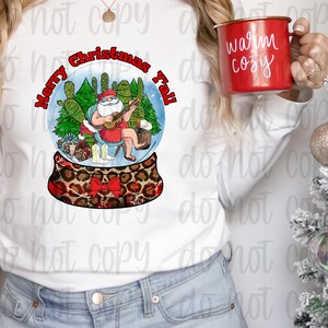 May include: A white long-sleeved shirt with a snow globe design featuring a cartoon Santa Claus playing a guitar in a desert setting. The snow globe is surrounded by the text "Merry Christmas Y'all".