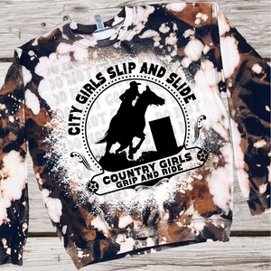 May include: A brown and white tie-dye sweatshirt with a black and white graphic design. The graphic features a silhouette of a person riding a horse over a barrel with the text "City Girls Slip and Slide Country Girls Grip and Ride".