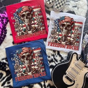 May include: Three t-shirts in red, white, and blue, each featuring a skull with an American flag bandana and peace signs. The text "FREEDOM ROCKS" is printed below the skull. A black and white guitar and checkered shoes are also visible.