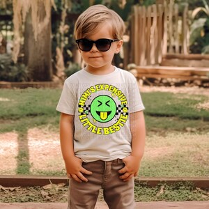 May include: A young child wearing a white t-shirt with a green and yellow graphic that says "Mimi's Expensive Little Bestie". The graphic features a cartoon smiley face with a tongue sticking out.