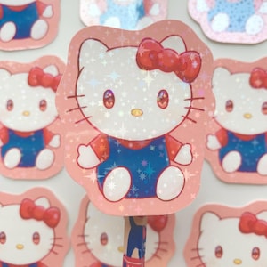 May include: Pink and holographic stickers featuring the cartoon character Hello Kitty. The stickers show Hello Kitty wearing a red bow and blue dungarees. The stickers are arranged around a pencil.